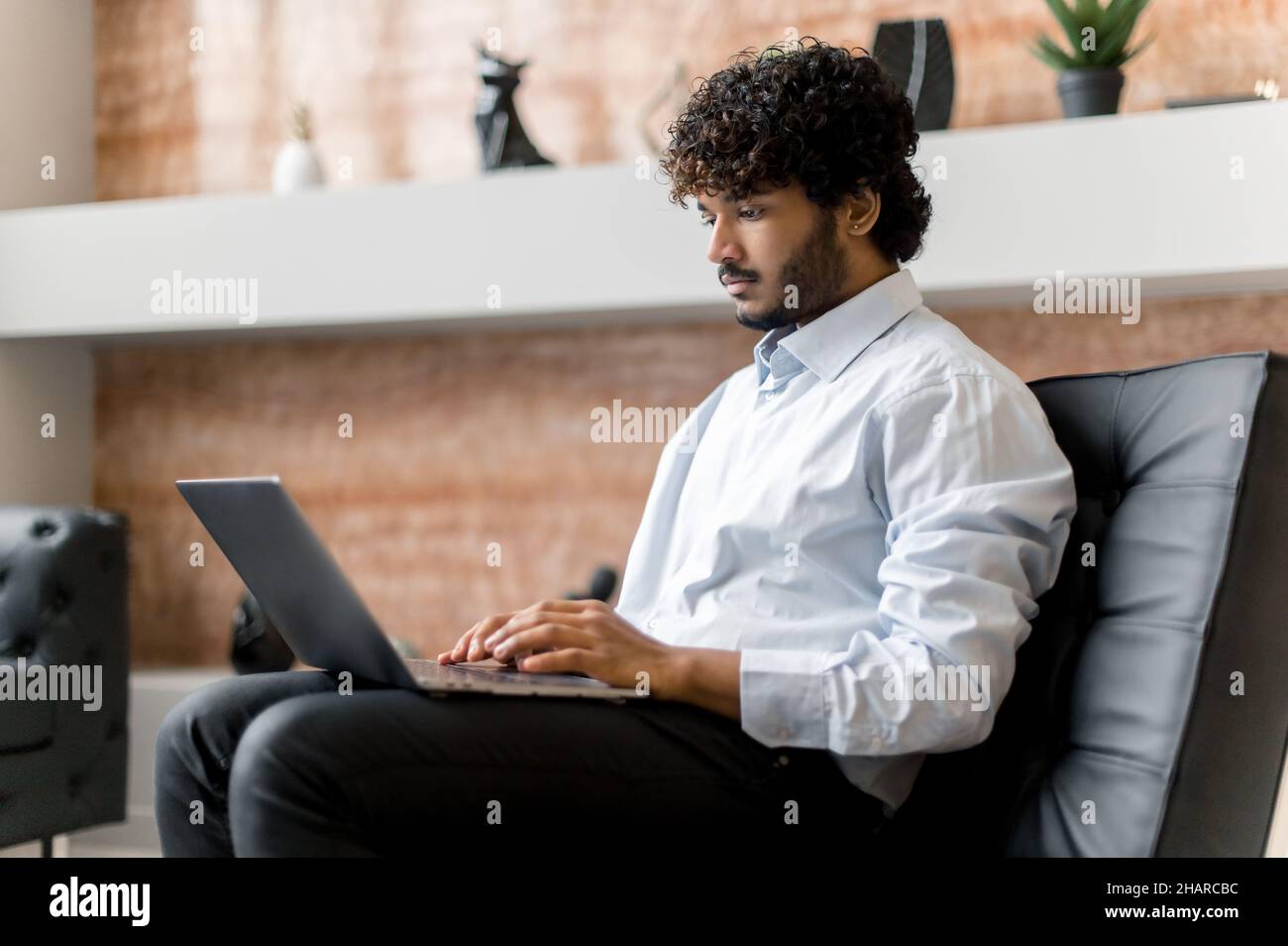Konzentrierter, ernsthafter, lockiger, indischer Mann mit lockigen Haaren, sitzend im Sessel, mit Laptop, mit Freunden, Kollegen oder Familie, Chatten in sozialen Medien, Beantworten von E-Mails Stockfoto