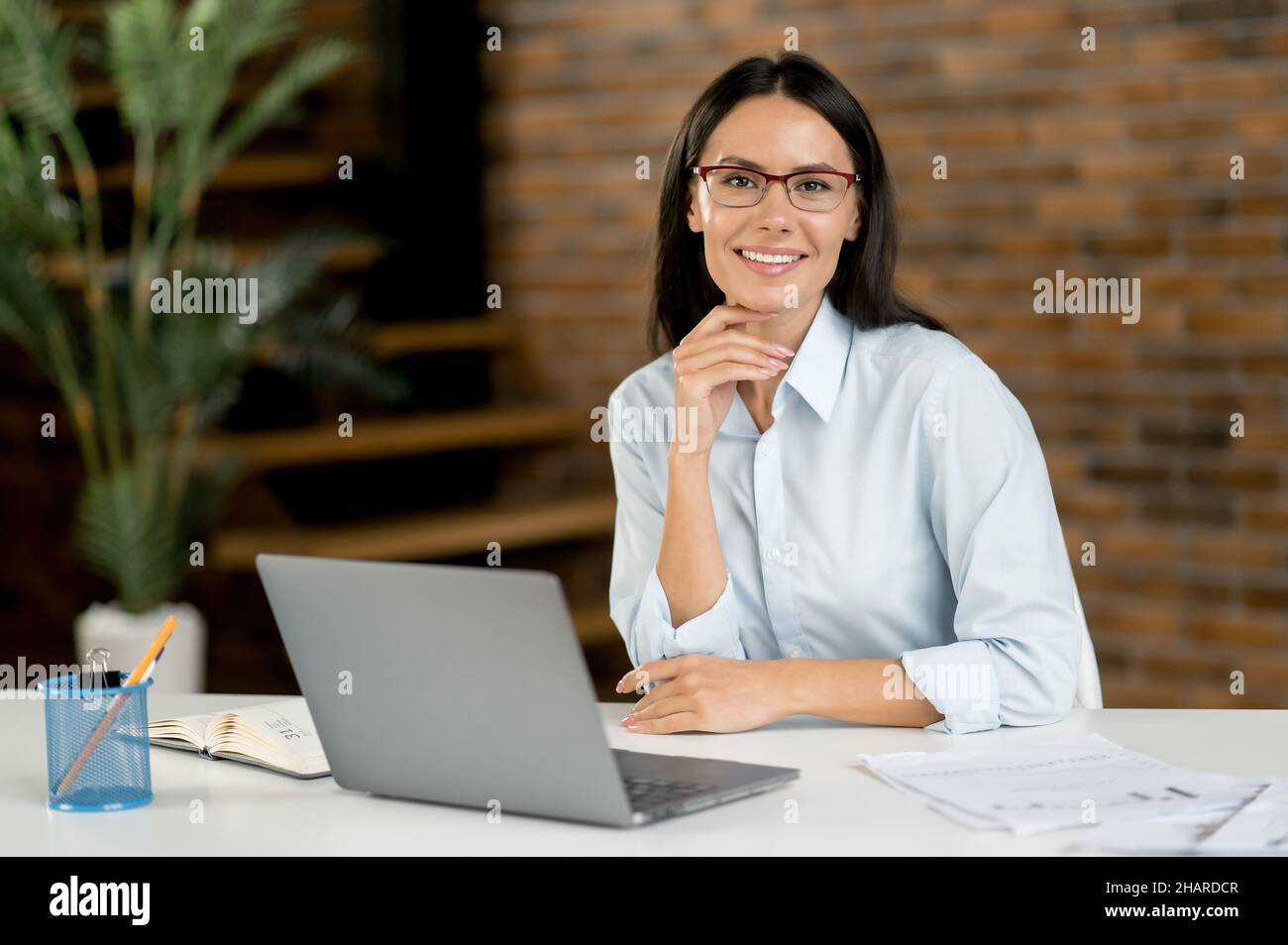 Smarte, selbstbewusste, erfolgreiche kaukasische Geschäftsfrau, geschäftsführerin oder Geschäftsführerin mit Brille, sitzt im Büro, mit einem Laptop, in stilvoller Kleidung, blickt auf die Kamera und lächelt Stockfoto