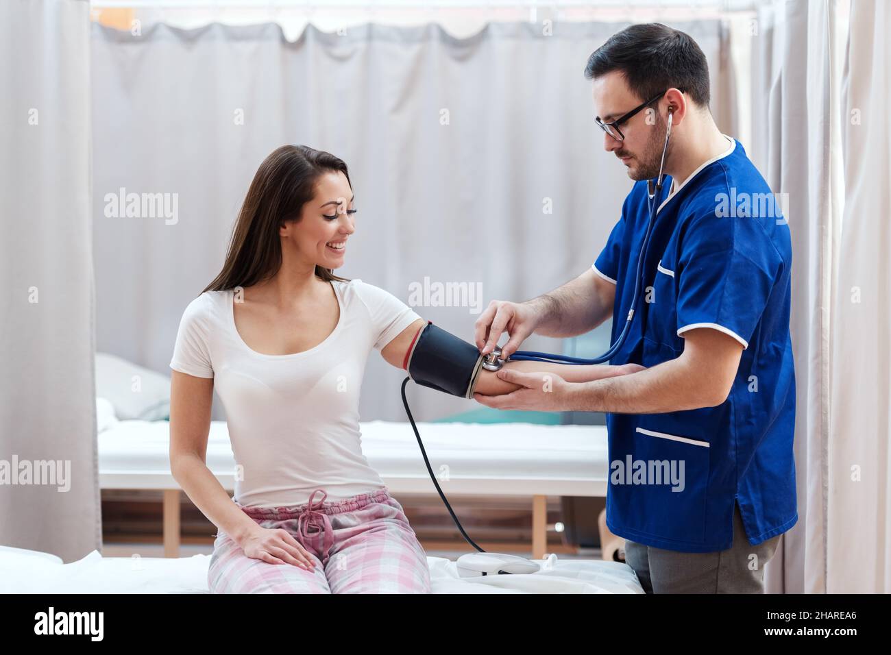 Medizinisches Personal steht und misst den Blutdruck, während sein Patient lächelt und auf dem Krankenhausbett sitzt. Stockfoto
