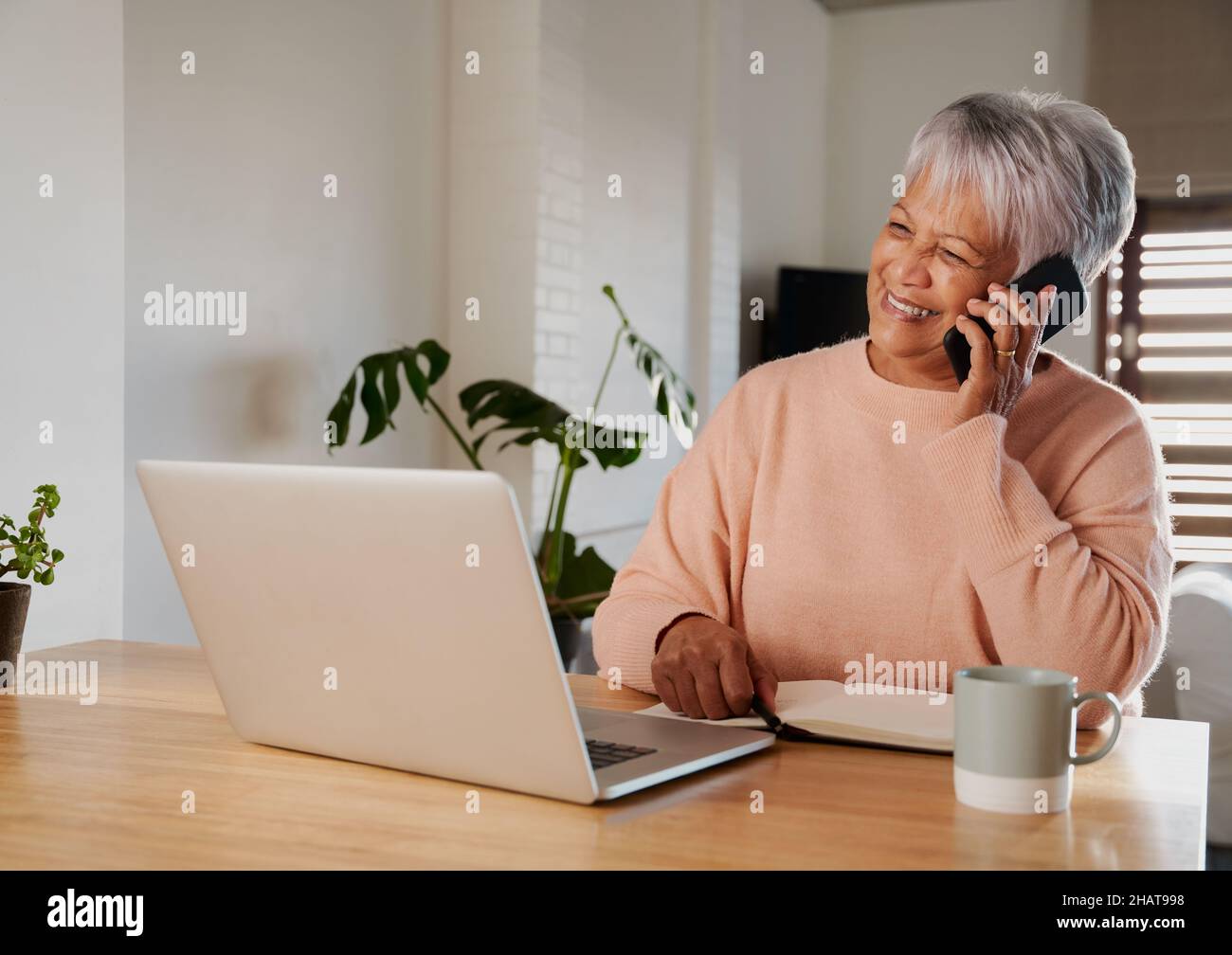 Multikulturelle ältere Frau, die vor dem Laptop an der Küchentischtheke sitzt, während sie mit der Familie telefoniert. Glücklicher Lebensstil im Ruhestand. Stockfoto