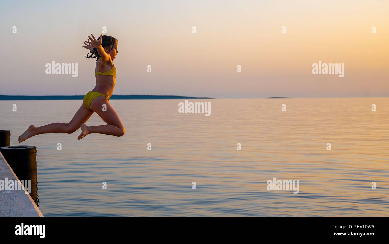 Mädchen, das bei Sonnenuntergang vom Pier im Wasser springt. Kinder springen im Meer in Silba, Kroatien. Stockfoto