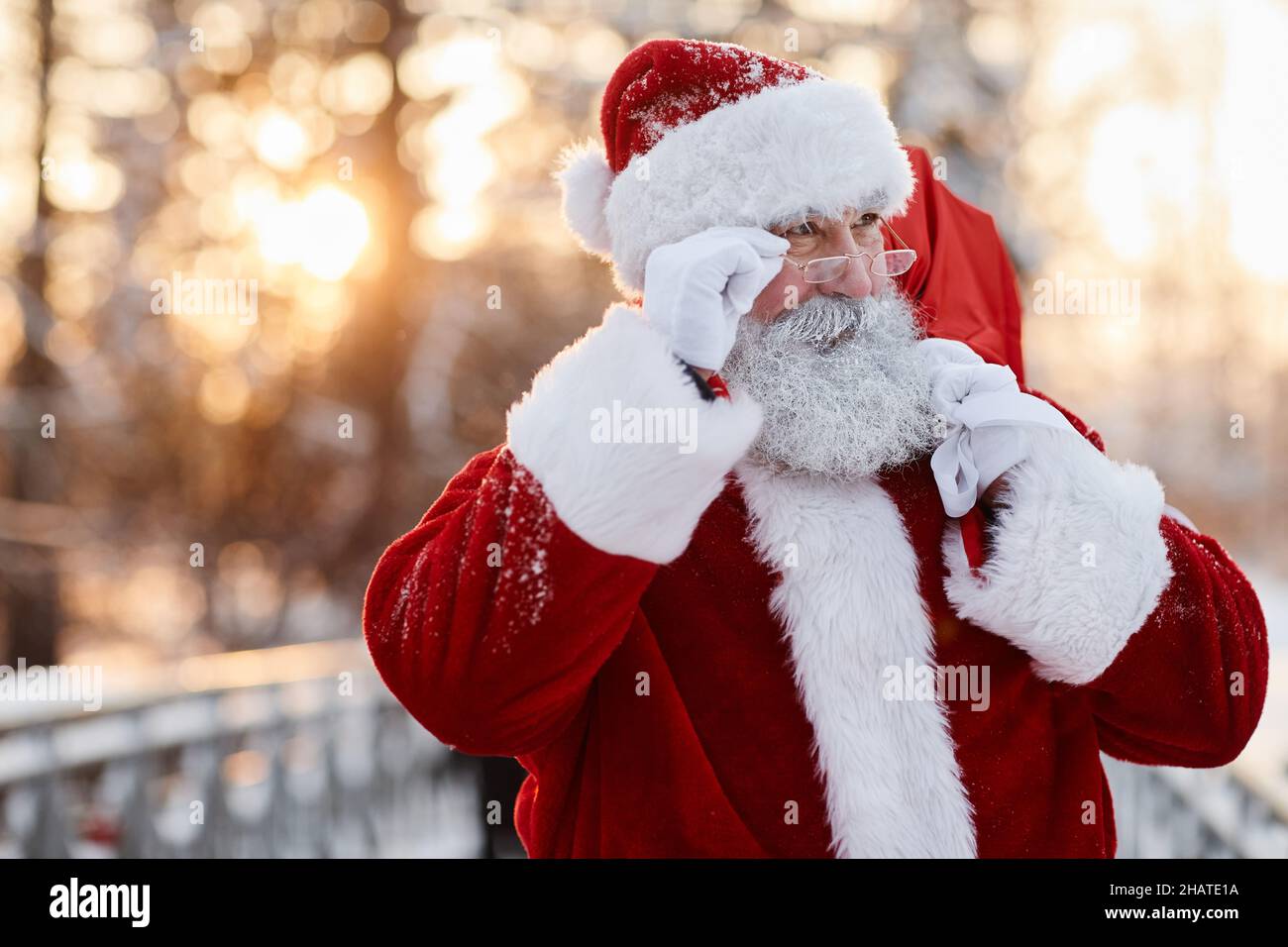 Horizontales, mittleres Nahaufnahme-Porträt des Weihnachtsmannes mit Brillen, der im Freien unterwegs ist und einen Sack mit Geschenken trägt, die wegschauen Stockfoto