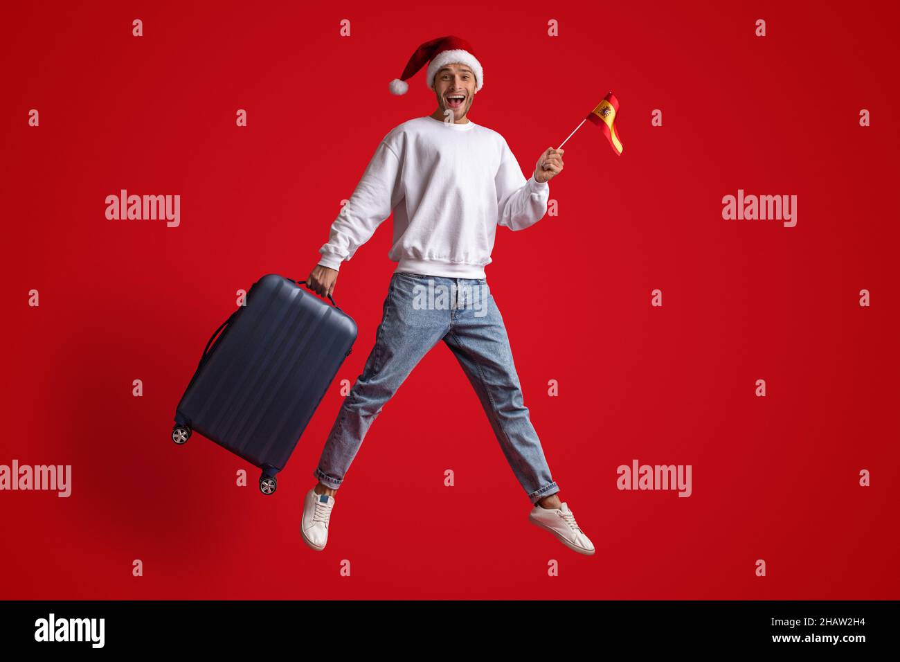 Glücklicher Männlicher Reisender Trägt Den Weihnachtsmütze, Der Mit Der Spanischen Flagge Und Dem Koffer Springt Stockfoto