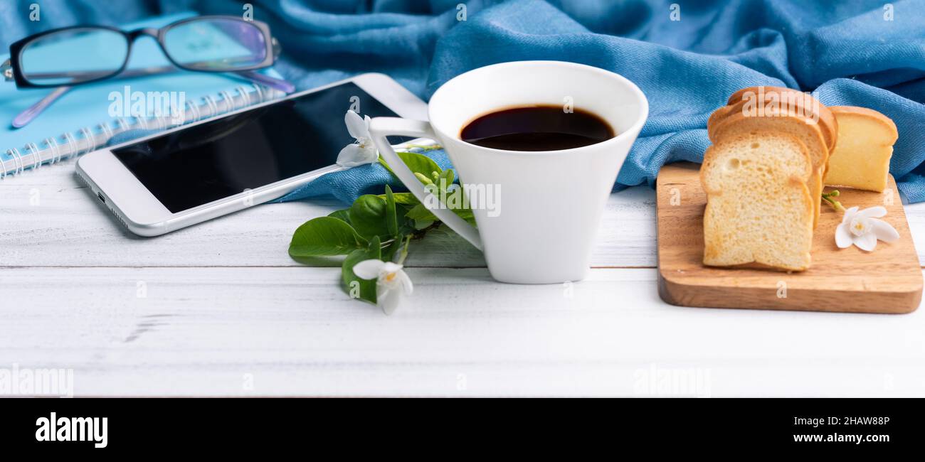 Frühstücksset mit Kaffeetasse, knusprigem Butterbrot, Mobiltelefon, Augenbrille und blauem Notizbuch auf weißem Holztisch Stockfoto