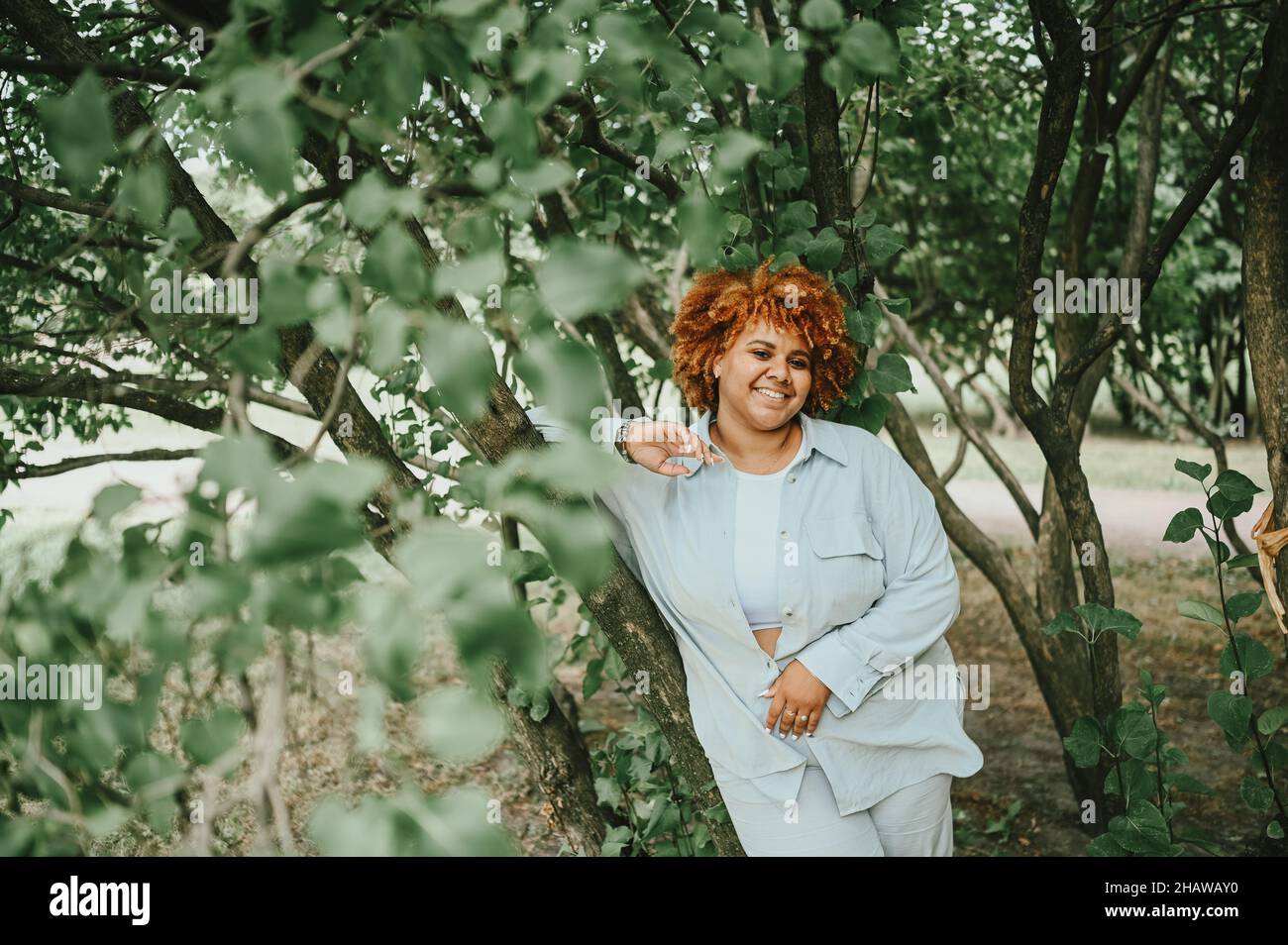 Fashion Portrait von jungen natürlich schönen plus size afroamerikanische Frau in hellen Hosenanzug mit afroroten Haaren posiert in Natur Parkland in grünem Laub. Vielfalt und körperpositives Konzept Stockfoto