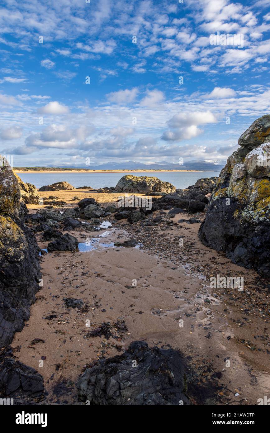 Ein felsiger Strand bei Ebbe auf Ynys Llanddwyn mit den Snowdonia-Bergen über der Bucht, Isle of Anglesey, Nordwales Stockfoto