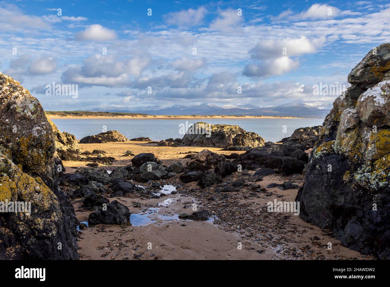 Ein felsiger Strand bei Ebbe auf Ynys Llanddwyn mit den Snowdonia-Bergen über der Bucht, Isle of Anglesey, Nordwales Stockfoto