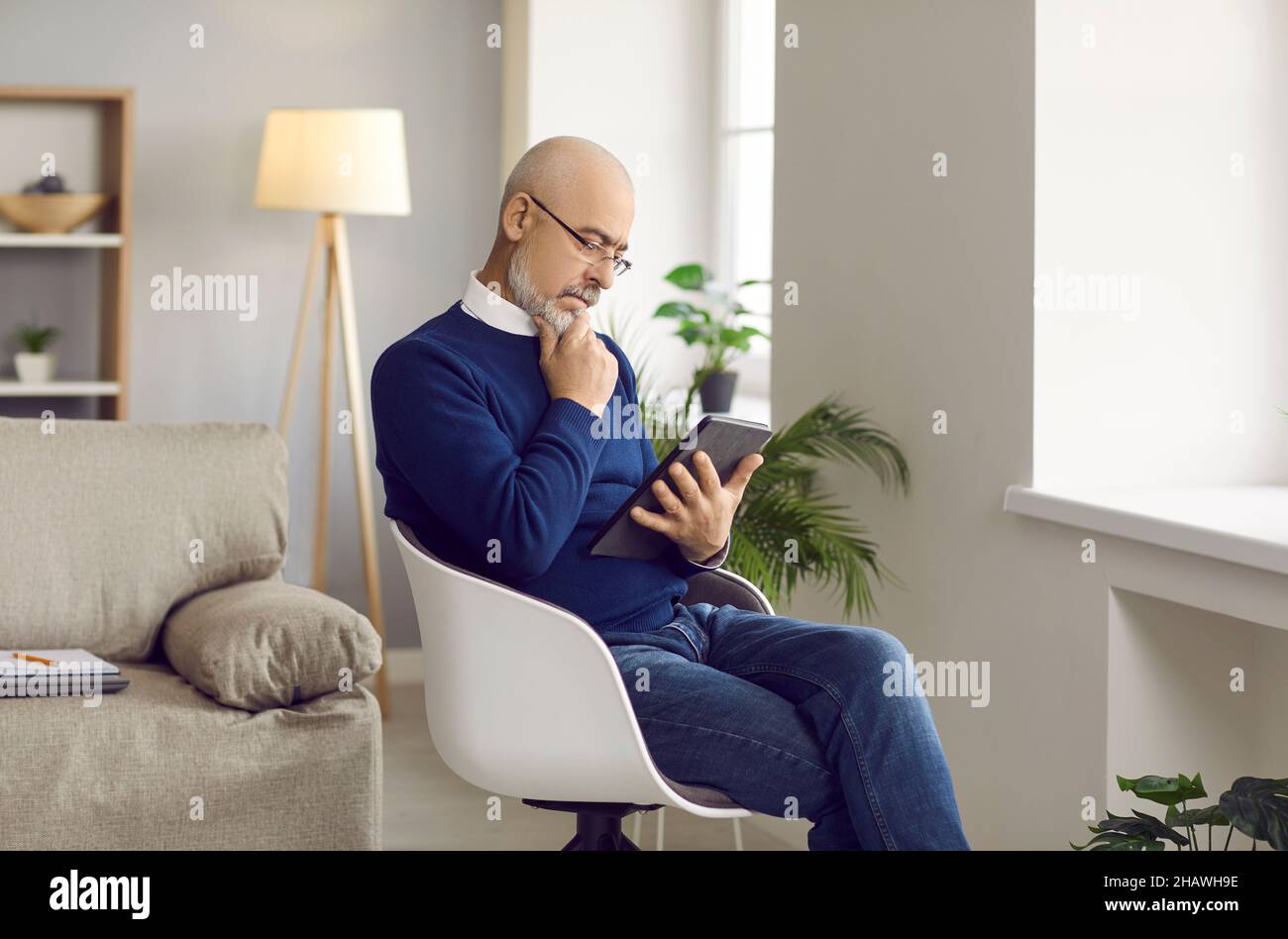 Älterer Mann, der zu Hause auf einem Stuhl sitzt, ein Tablet hält, ein eBook liest und nachdenkt Stockfoto