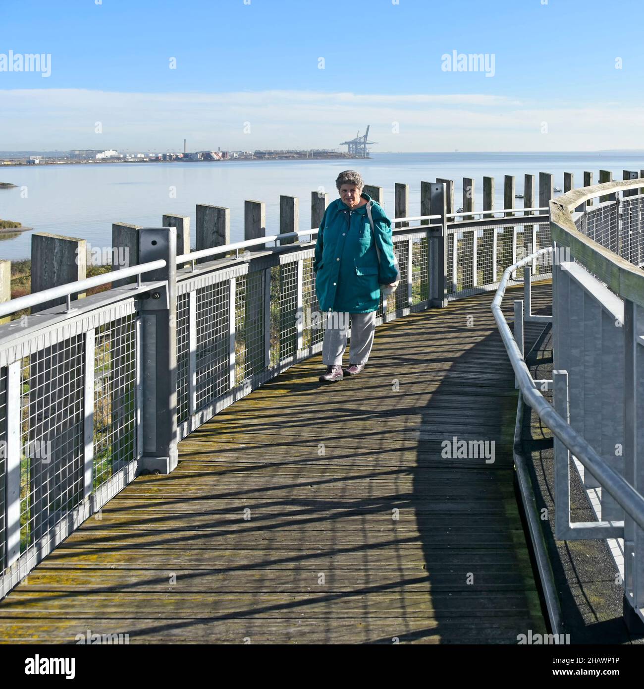 Ältere Frau, die auf dem Dach auf der Aussichtsplattform des Essex Wildlife Trust im Besucherzentrum des Thameside Nature Reserve Parks spazieren geht, mit Blick auf die Themse-Mündung in Großbritannien Stockfoto