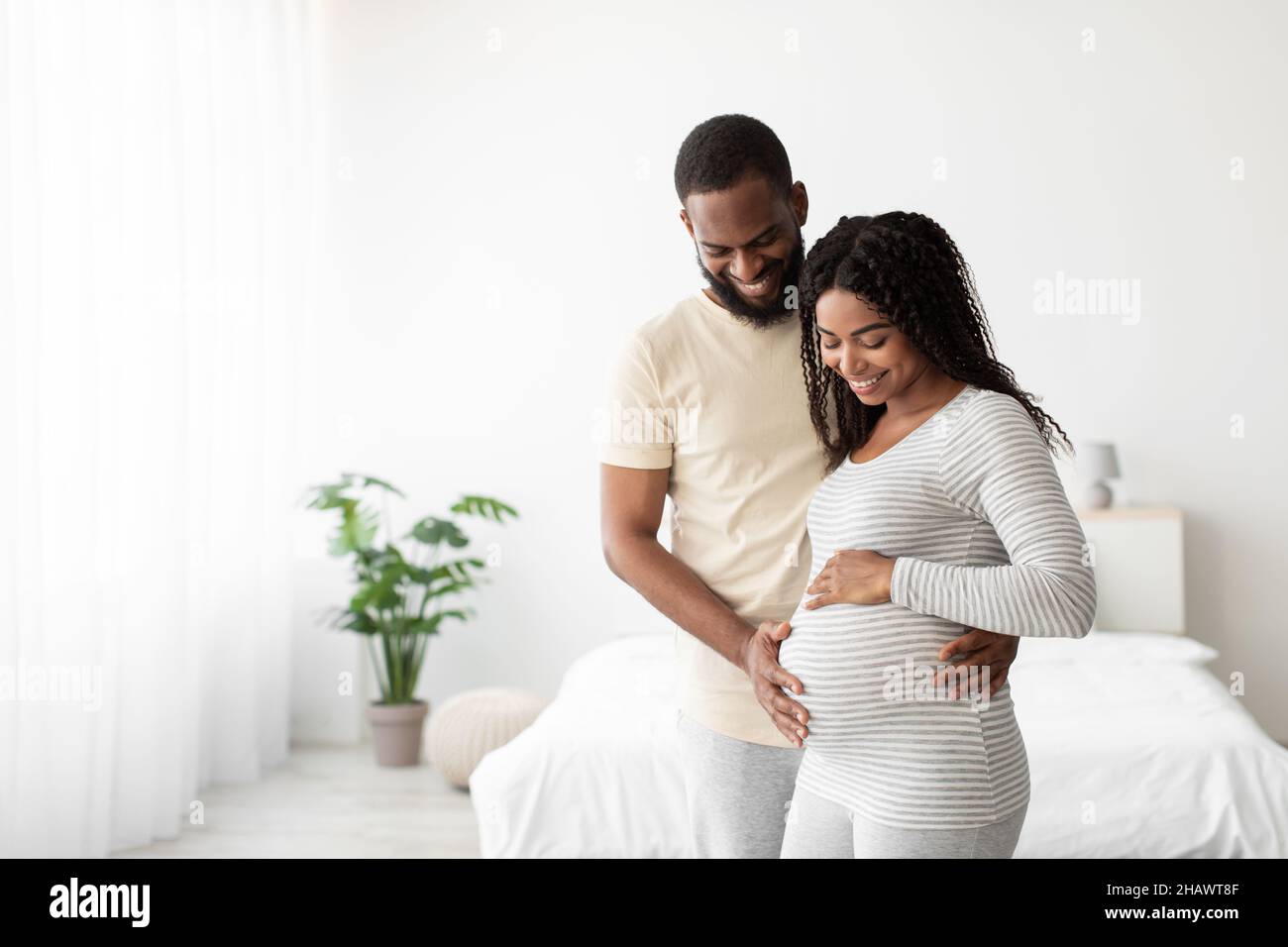 Lächelnder junger afroamerikanischer Ehemann umarmt die Schwangeren und berührt den Bauch im minimalistischen Schlafzimmer Stockfoto