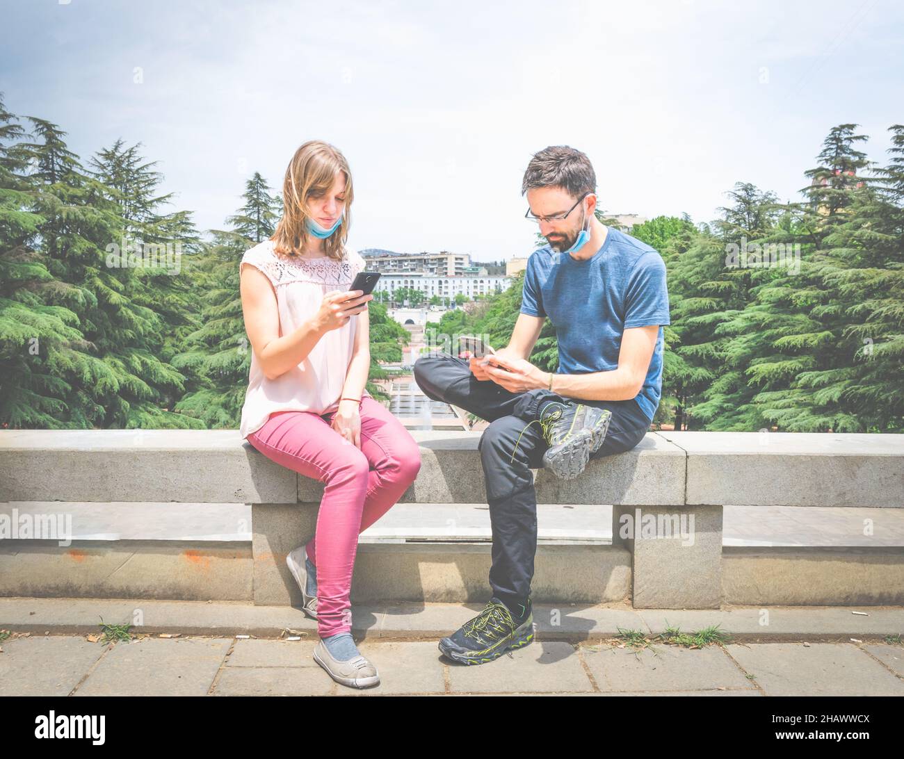 Zwei junge Menschen mit Masken sitzen auf den Zementblöcken in einem Park und schauen auf ihre Telefone. Soziale Medien und Sozialisierung während Pandemien. Stockfoto