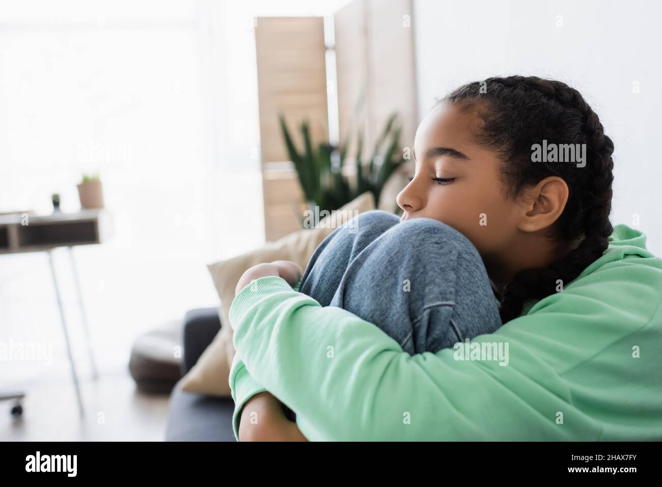 Verärgerte afroamerikanische Teenager-Mädchen umarmte Knie, während sie zu Hause mit geschlossenen Augen saß Stockfoto