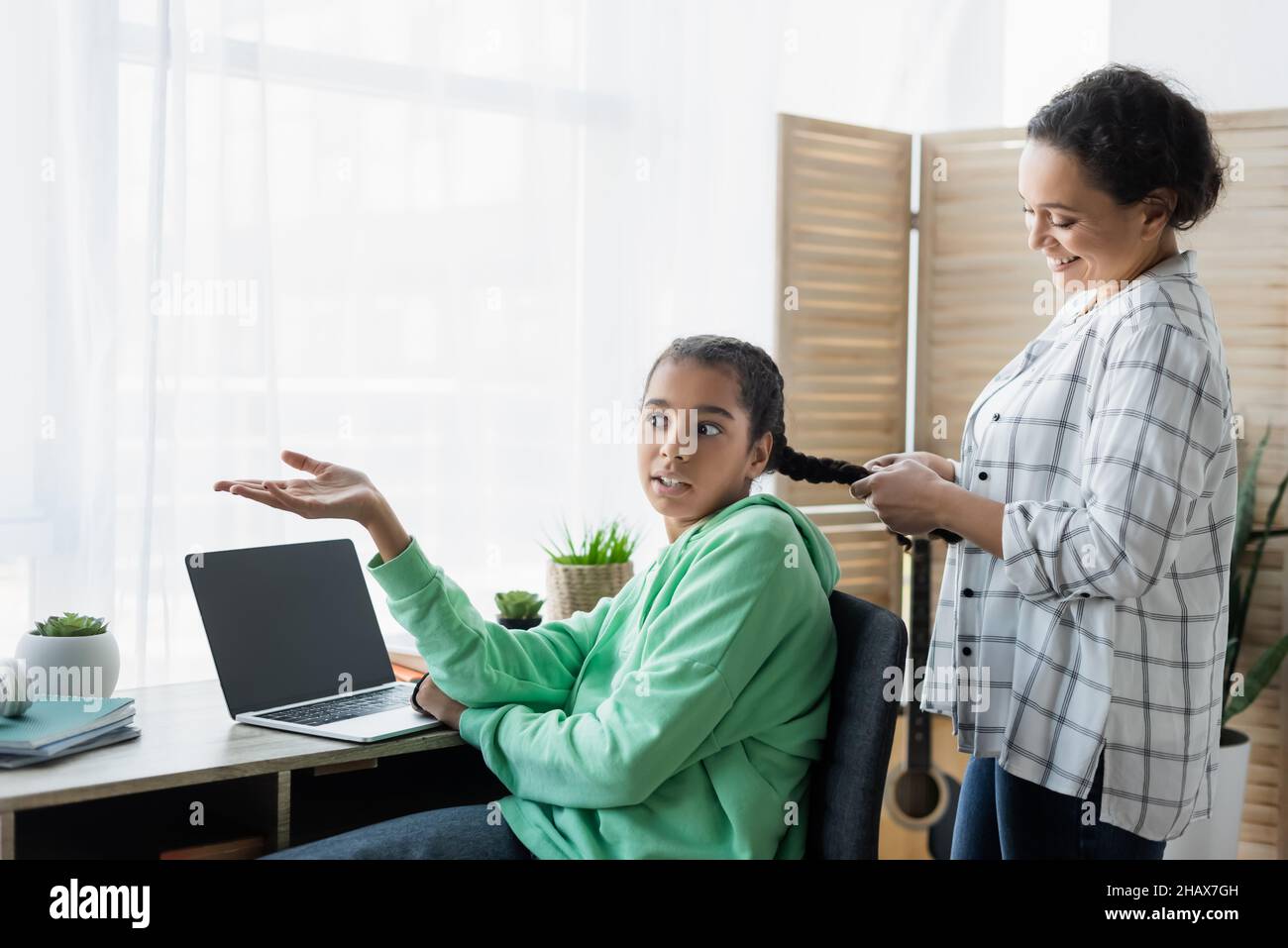 afroamerikanisches Mädchen zeigt auf Laptop mit leerem Bildschirm, während Mutter ihre Haare flechtet Stockfoto