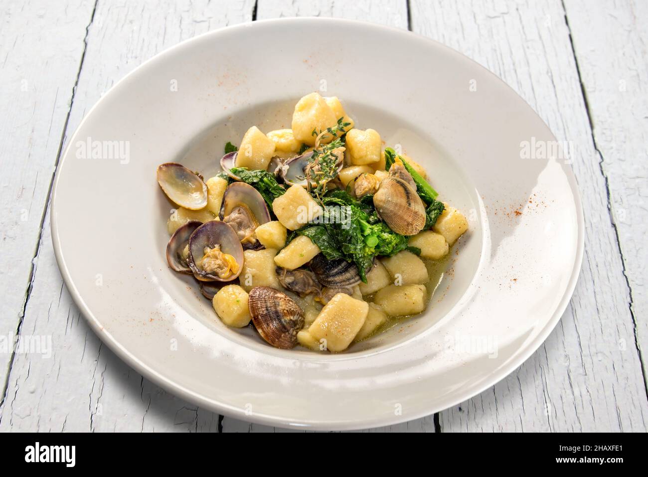 Kartoffelknödel-Gnocchi mit Venusmuscheln und Rübengemüse in weißem Teller auf weißem Holztisch Stockfoto