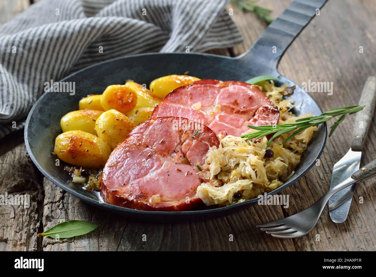 Deutsche Küche – geräucherter Schweinehalk mit Sauerkraut und gebratenen Babykartoffeln, die in einer eisernen Pfanne auf einem alten Holztisch serviert werden Stockfoto