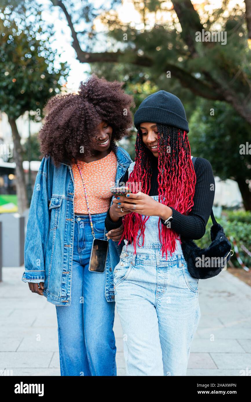 Junge ethnische Freundinnen mit Afro-Frisuren konzentrieren sich, während sie gemeinsam auf einer verschwommenen Straße in der Stadt stehen und Mobiltelefone durchsuchen Stockfoto