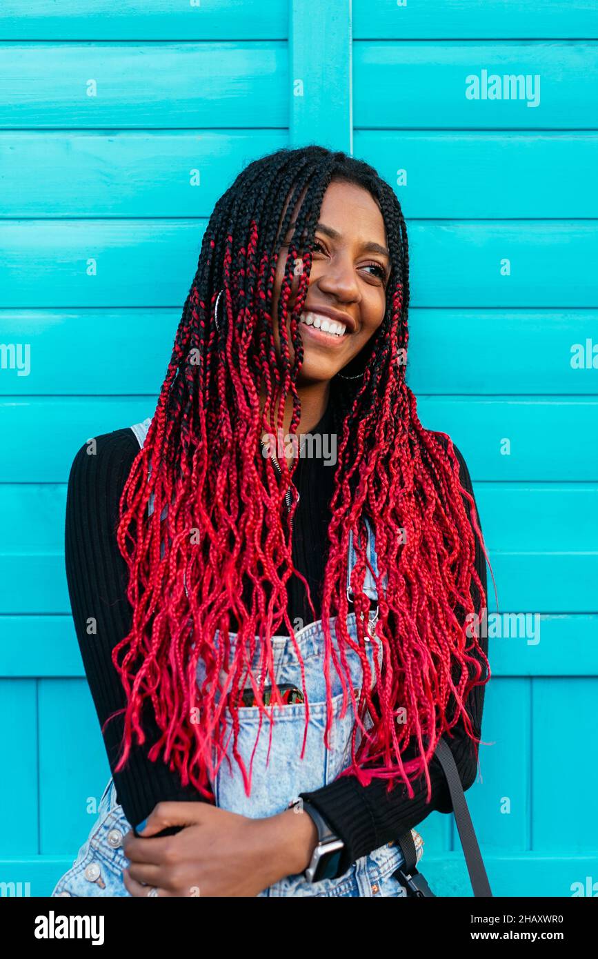 Fröhliche ethnische Hündin mit roter Afro-Frisur, die an der blauen Straßenwand steht und wegschaut Stockfoto