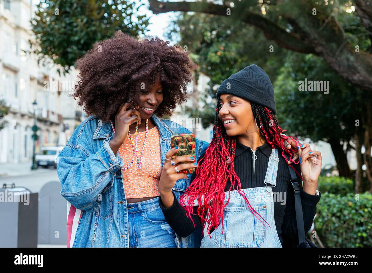 Junge ethnische Freundinnen mit Afro-Frisuren konzentrieren sich, während sie gemeinsam auf einer verschwommenen Straße in der Stadt stehen und Mobiltelefone durchsuchen Stockfoto