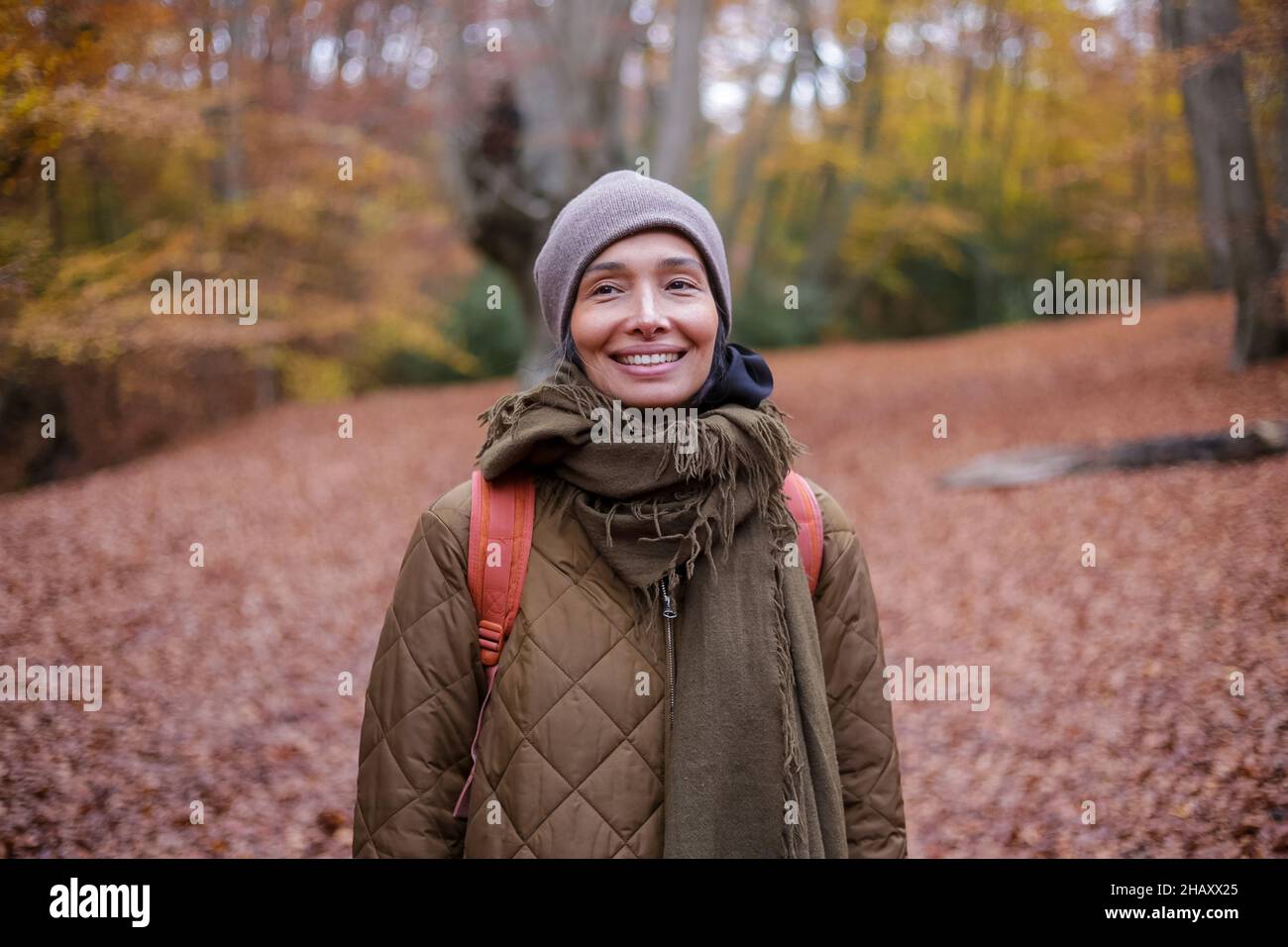 Fröhliche, ethnische Frau in Oberbekleidung und Hut lächelnd und schauend auf die Kamera, während sie am Wochenende inmitten von Bäumen im Herbstwald steht Stockfoto