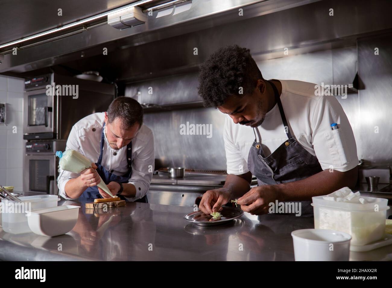 Konzentrierter schwarzer männlicher Koch kocht Gericht in der Nähe von Kollegen, der in der Küche des Restaurants die Creme aus dem Gebäckbeutel auf die Vorspeise drückt Stockfoto