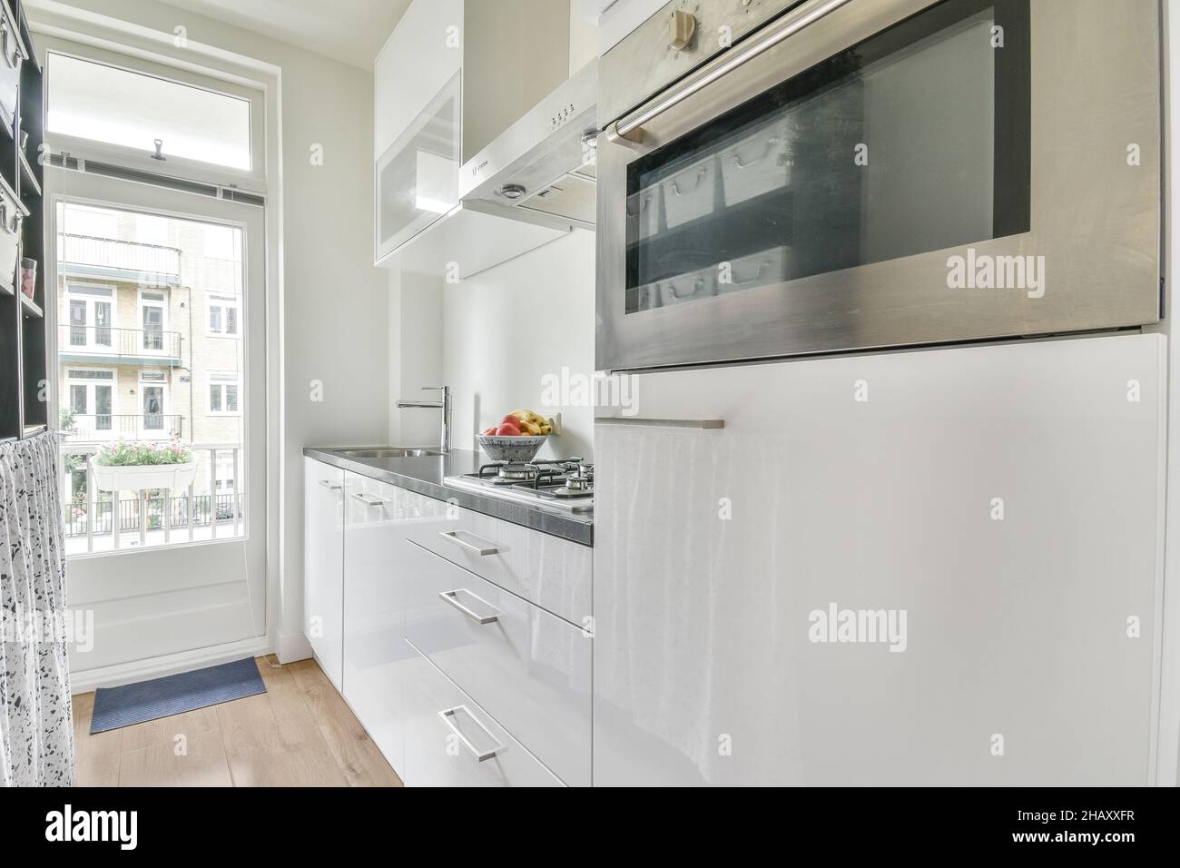 Interieur der modernen hellen schmalen Küche mit weißen Schränken mit eingebautem Ofen und Zähler mit Waschbecken und Herd auf Parkettboden in der Nähe eingerichtet Stockfoto