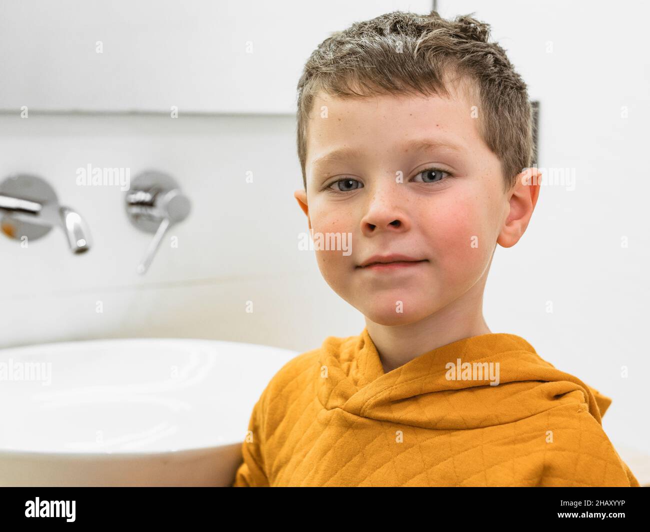 Süßer Junge in legerem Outfit, der während der täglichen Hygiene in der Nähe eines weißen Waschbeckens mit Wasserhahn im Badezimmer die Kamera anschaut Stockfoto