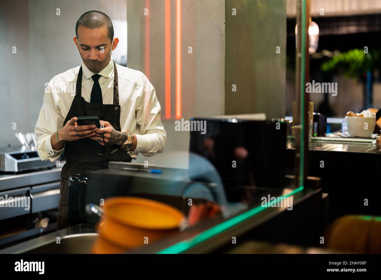 Durch Glas seriöse Kellner in Uniform und Maske zum Schutz vor COVID 19 Surfen Handy im Restaurant Stockfoto