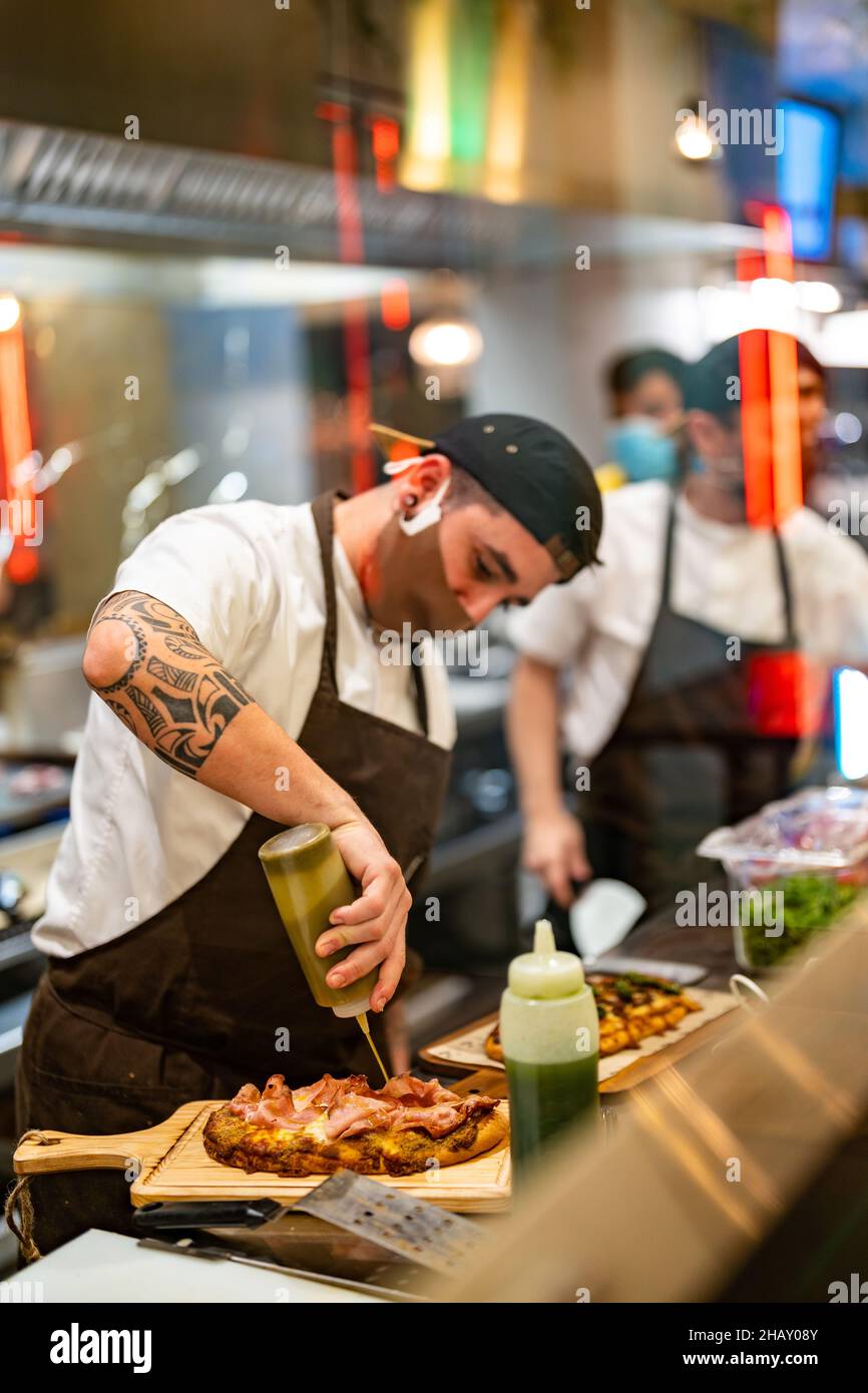 Durch ein Glas beschäftigten männlichen Koch in Maske Zugabe von Sauce aus der Flasche zu leckeren Pizza auf Holz Schneidebrett während der Arbeit in der offenen Küche des Restaurants du Stockfoto