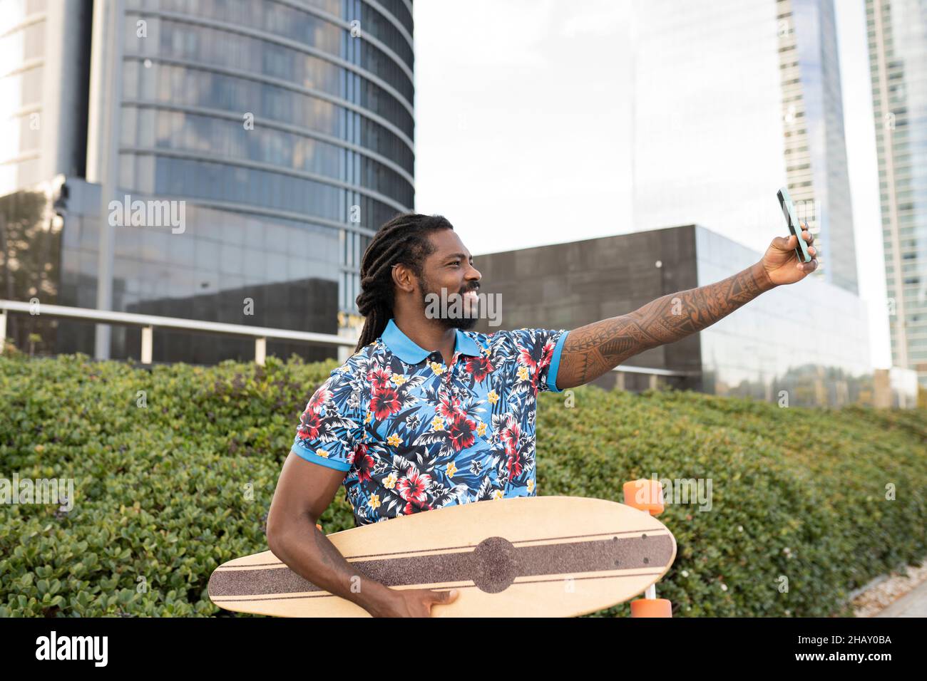 Seitenansicht des entzückten afroamerikanischen Mannes in legerer Kleidung mit Longboard, der auf dem Smartphone Selbstporträt aufnahm, während er auf der Straße der Stadt stand Stockfoto