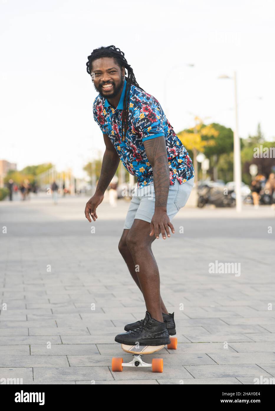 Ein ganzer Körper von fröhlichem, sportlichem afroamerikanischem Mann in lässiger Kleidung, der auf einem befestigten Gehweg auf der Straße der Stadt auf einem Longboard reitet Stockfoto