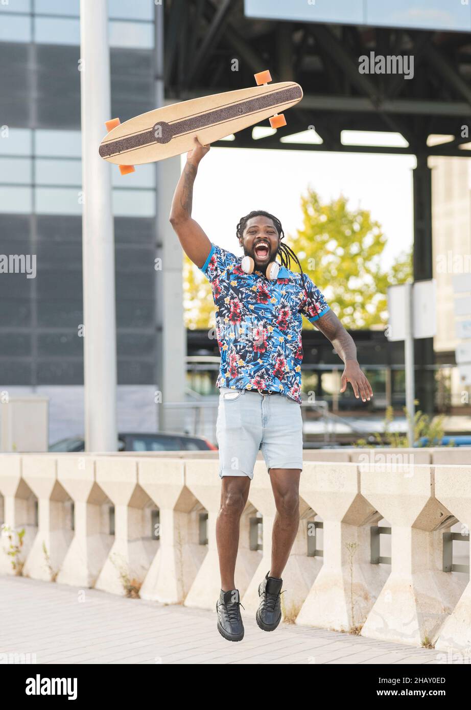 Aufgeregter afroamerikanischer Mann mit kabellosen Kopfhörern, der mit Longboard in der Hand auf einem gepflasterten Gehweg in der Stadt hüpft Stockfoto