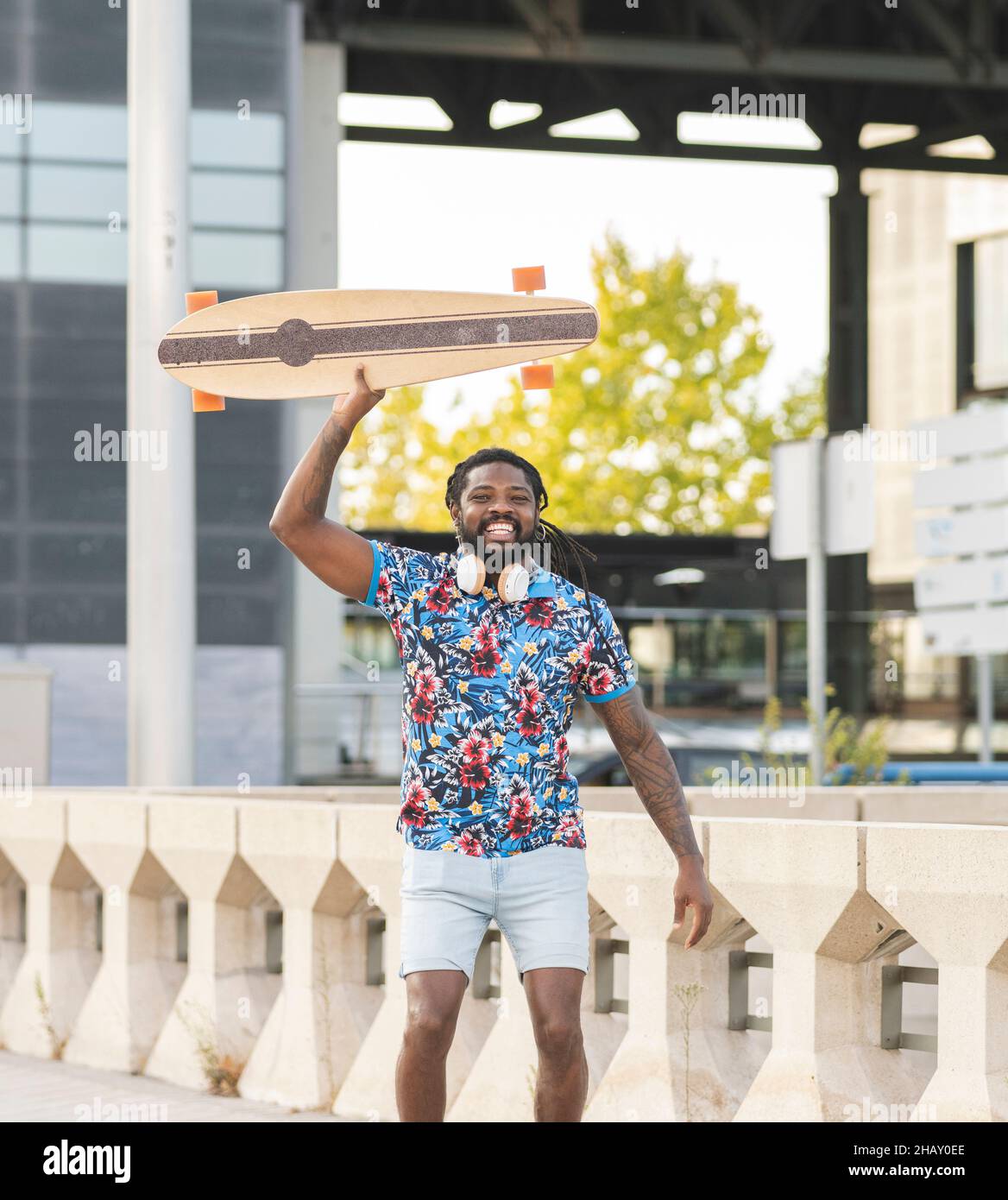 Aufgeregter afroamerikanischer Mann mit kabellosen Kopfhörern, der mit Longboard in der Hand auf einem gepflasterten Gehweg in der Stadt hüpft Stockfoto