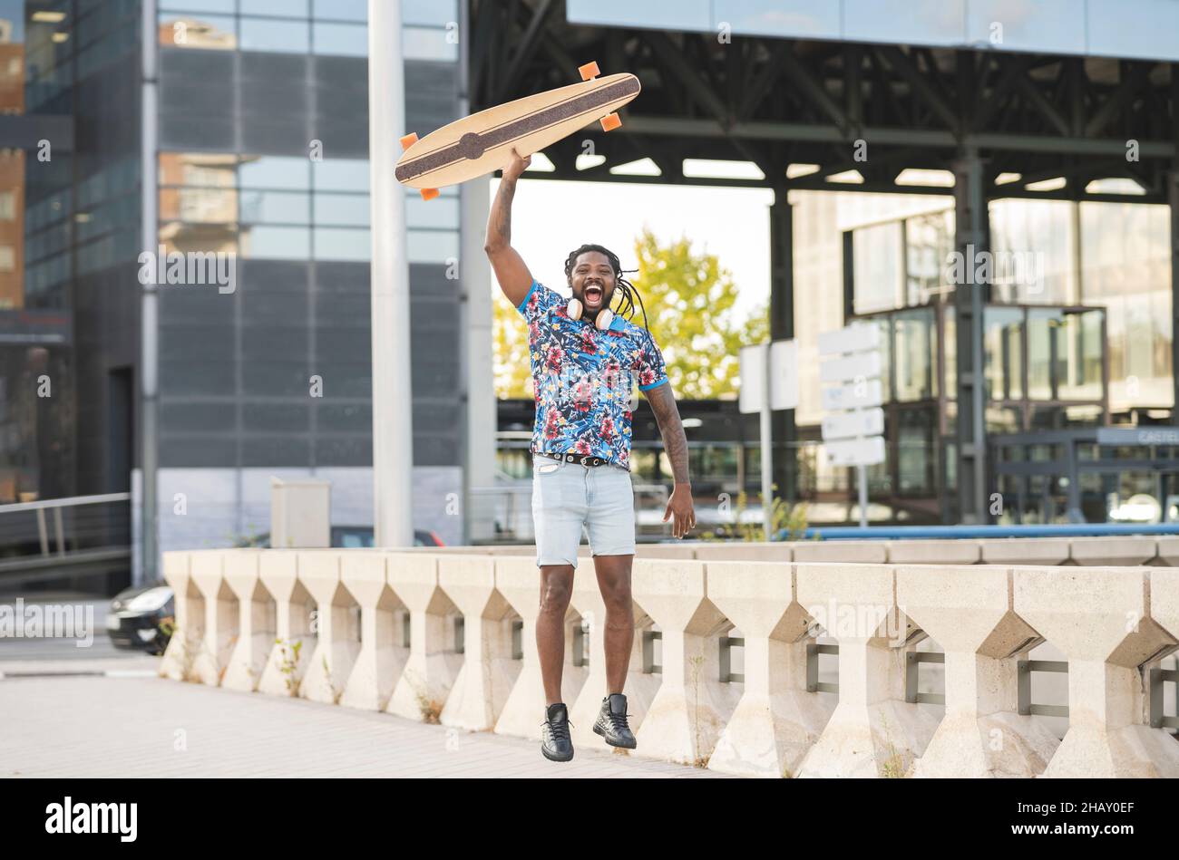 Aufgeregter afroamerikanischer Mann mit kabellosen Kopfhörern, der mit Longboard in der Hand auf einem gepflasterten Gehweg in der Stadt hüpft Stockfoto