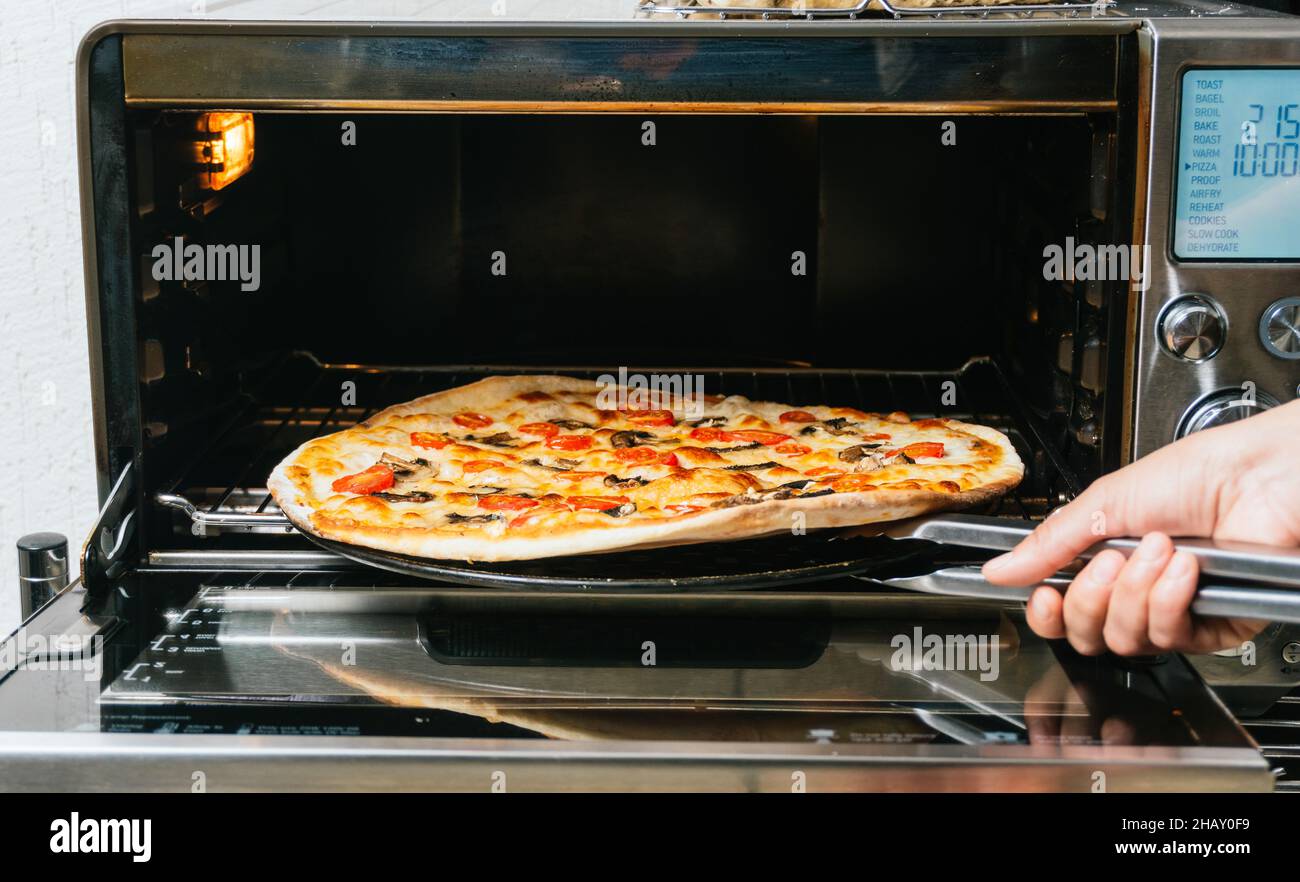 Crop anonyme Köchin Entfernen von heiß gebackenen Pizza mit Tomaten und Pilzen und Käse aus dem kleinen Ofen Stockfoto