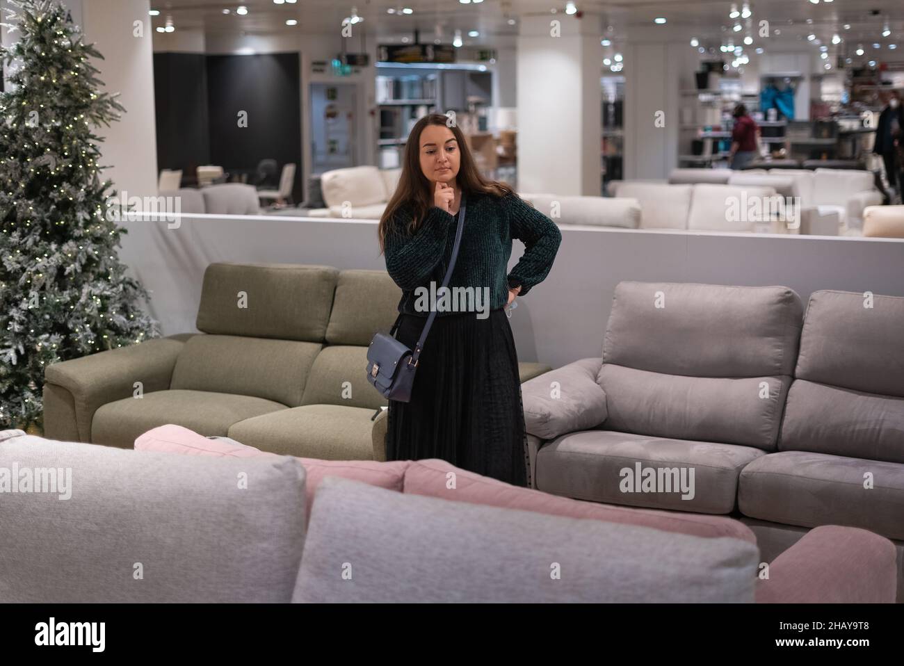Weibliche Klientin berührt Kinn und denkt, während sie eine Couch im modernen Möbelgeschäft auswählt Stockfoto