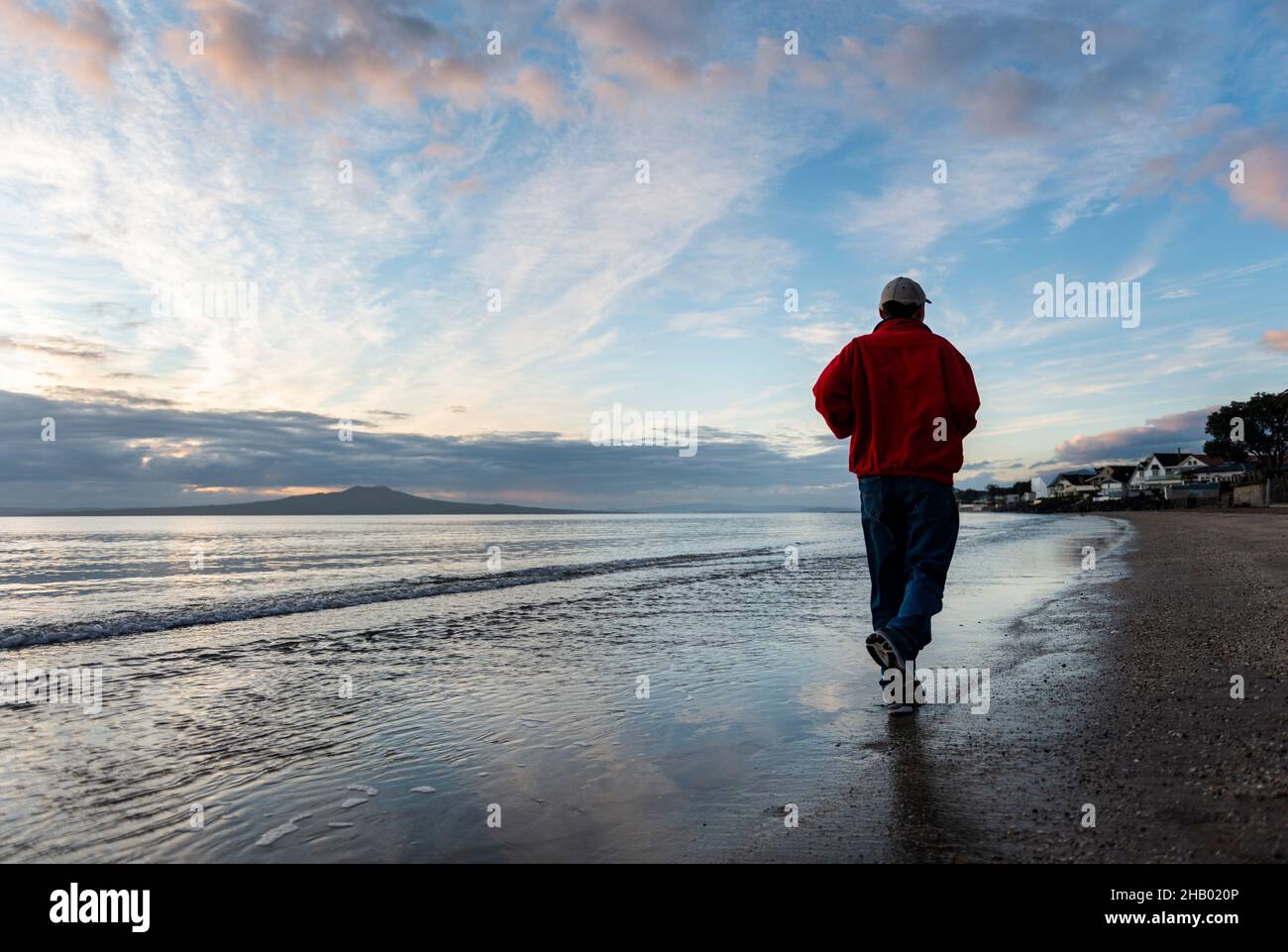 Mann, der bei Sonnenaufgang am Strand von Milford mit Rangitoto Island in der Ferne, Auckland, läuft. Stockfoto