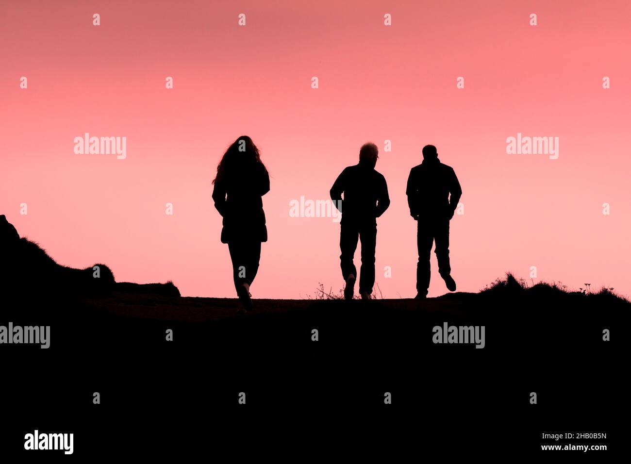 Drei Menschen, die am Ende des Tages in Newquay in Cornwall den Küstenpfad entlang wanderten, schillerten gegen einen bunten Himmel. Stockfoto