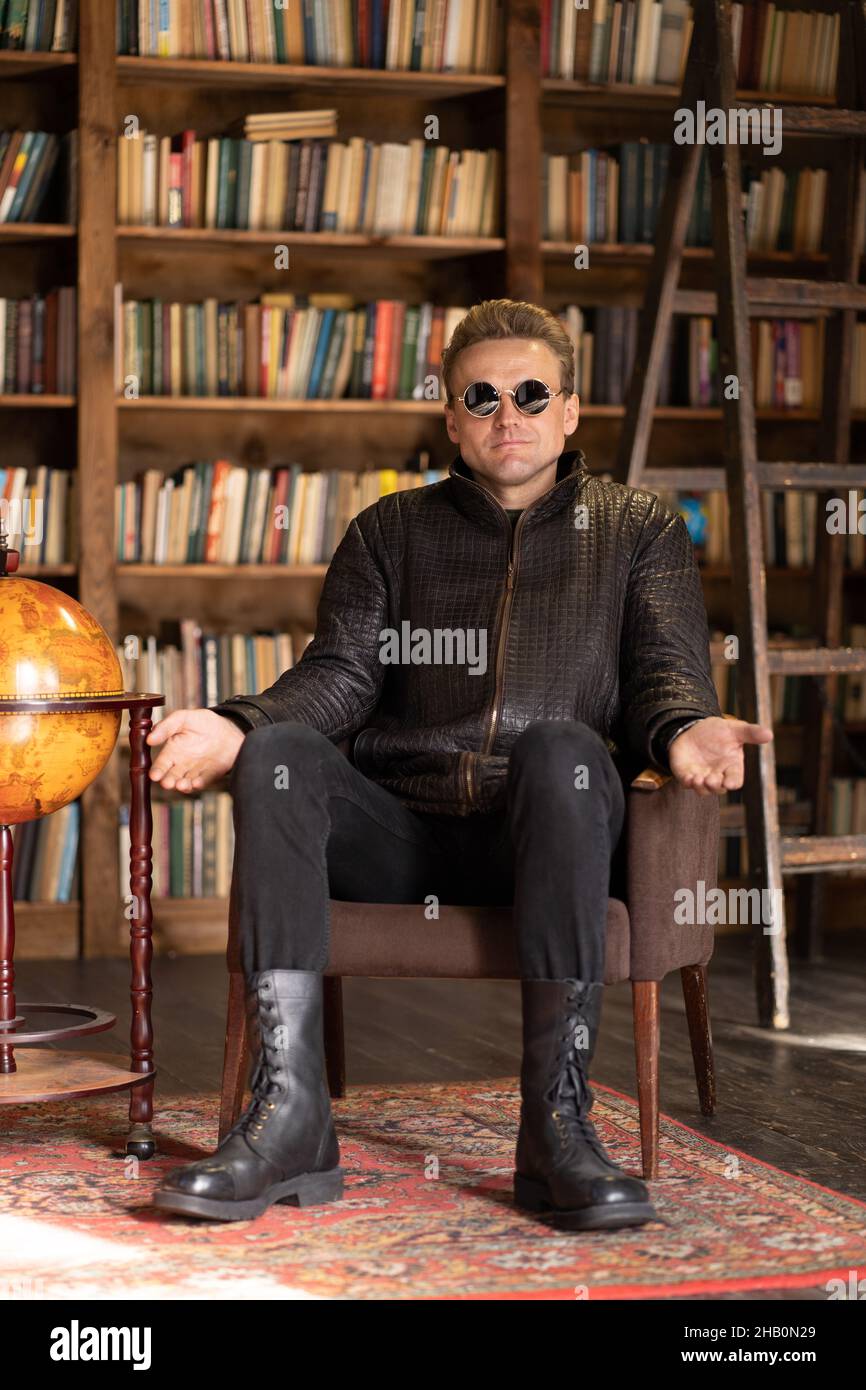 Männer in Hipster Lederjacke und stilvolle Sonnenbrille sitzt in einem Retro-Sessel mit offenen Händen in Loft Studio . Bücherregale Hintergrund. Nahaufnahme . Hochwertige Fotos Stockfoto