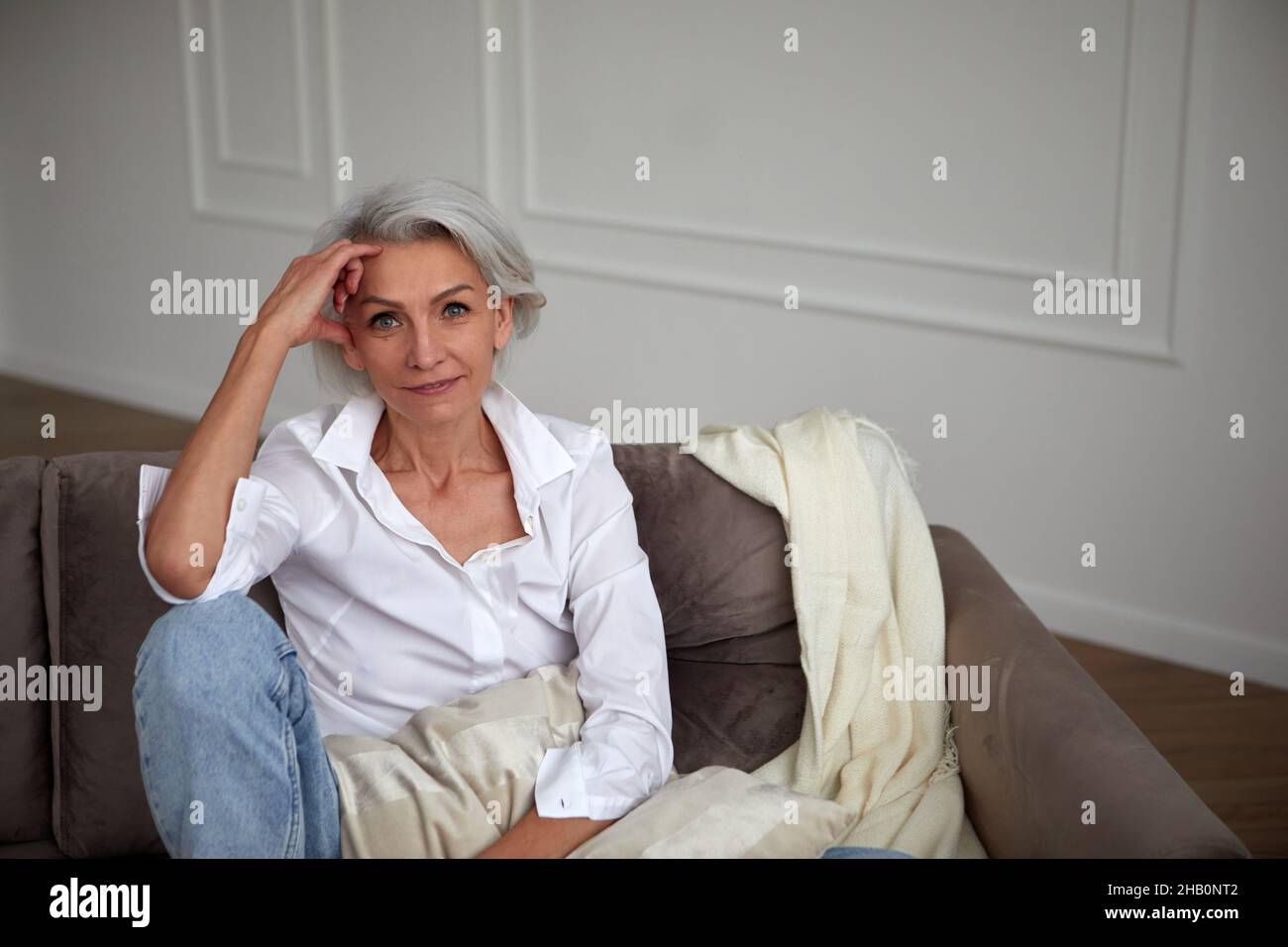 Lächelnde, stilvolle, reife Frau mit grauen Haaren, die auf dem Sofa sitzt und sich an der Hand lehnt, während sie im Wohnzimmer chillt und auf die Kamera schaut Stockfoto