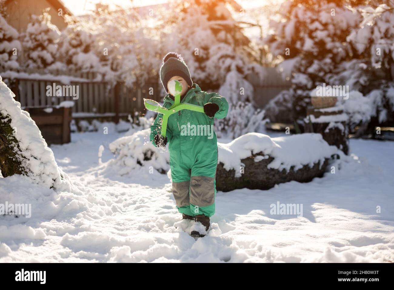 Junge spielt im Winter mit Schnee. Kleines Kind in grüner Jacke und gestricktem Hut machen an Weihnachten Schneebälle in der Nähe des Hauses. Kinder spielen und Spaß haben im verschneiten Garten Stockfoto