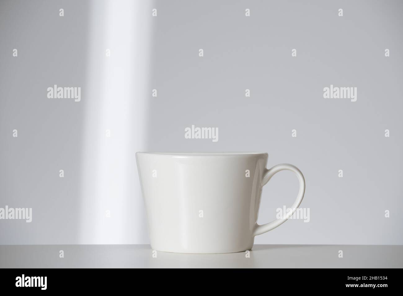 Weiße Tasse auf weißem Hintergrund mit Sonnenstrahl. Morgendliche Selbstpflege Routine Minimalismus und Vorbereitung. Vorderansicht Stockfoto