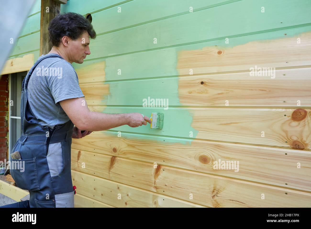 Der Mann in einem Arbeiteranzug malt die hölzerne Oberfläche der Wand grün Stockfoto