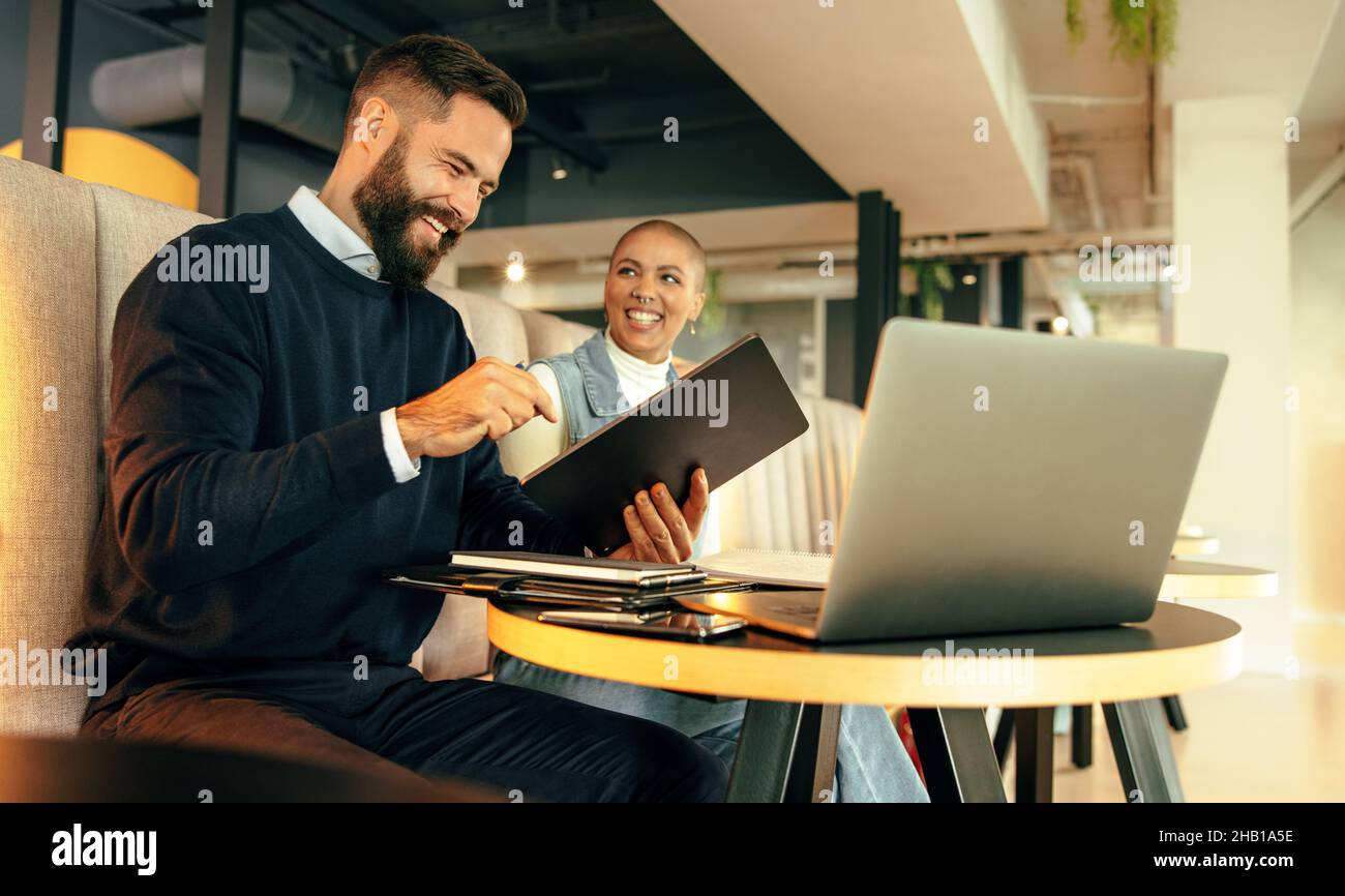 Zwei junge Geschäftsleute, die fröhlich lächeln, während sie in einer Bürolobby zusammenarbeiten. Zwei glückliche Unternehmer mit einem digitalen Tablet und einem Laptop in einem Stockfoto