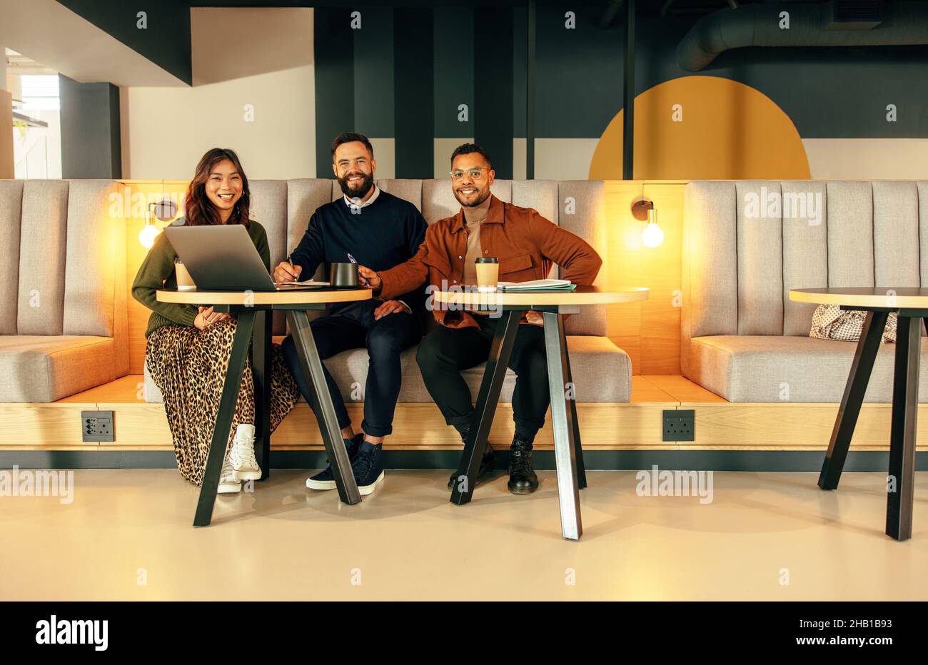 Glückliche Geschäftsleute, die in einer modernen Bürolobby arbeiten. Ein Team glücklicher Geschäftsleute, die auf der Kamera lächeln, während sie auf einer Couch sitzen. Gruppe von Unternehmungen Stockfoto