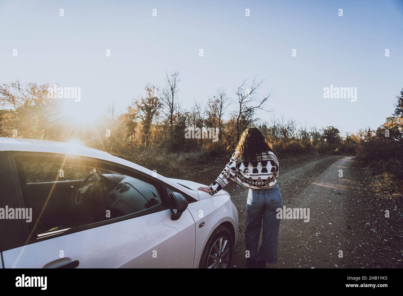 Frau neben einem Auto auf einer Straße zwischen Bäumen bei Sonnenuntergang Stockfoto