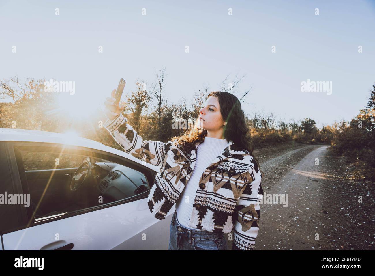 Frau, die auf das Handy neben einem Auto auf einer Straße zwischen Bäumen schaut Stockfoto