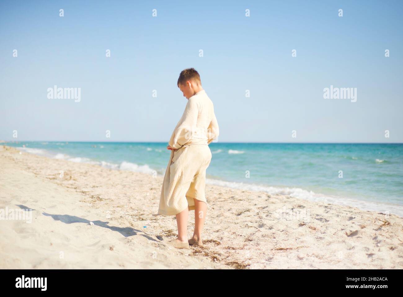 Ein barfuß-Junge in Musselin-Robe steht mit dem Rücken an der Küste. Türkisfarbenes Meer. Sand. Stockfoto