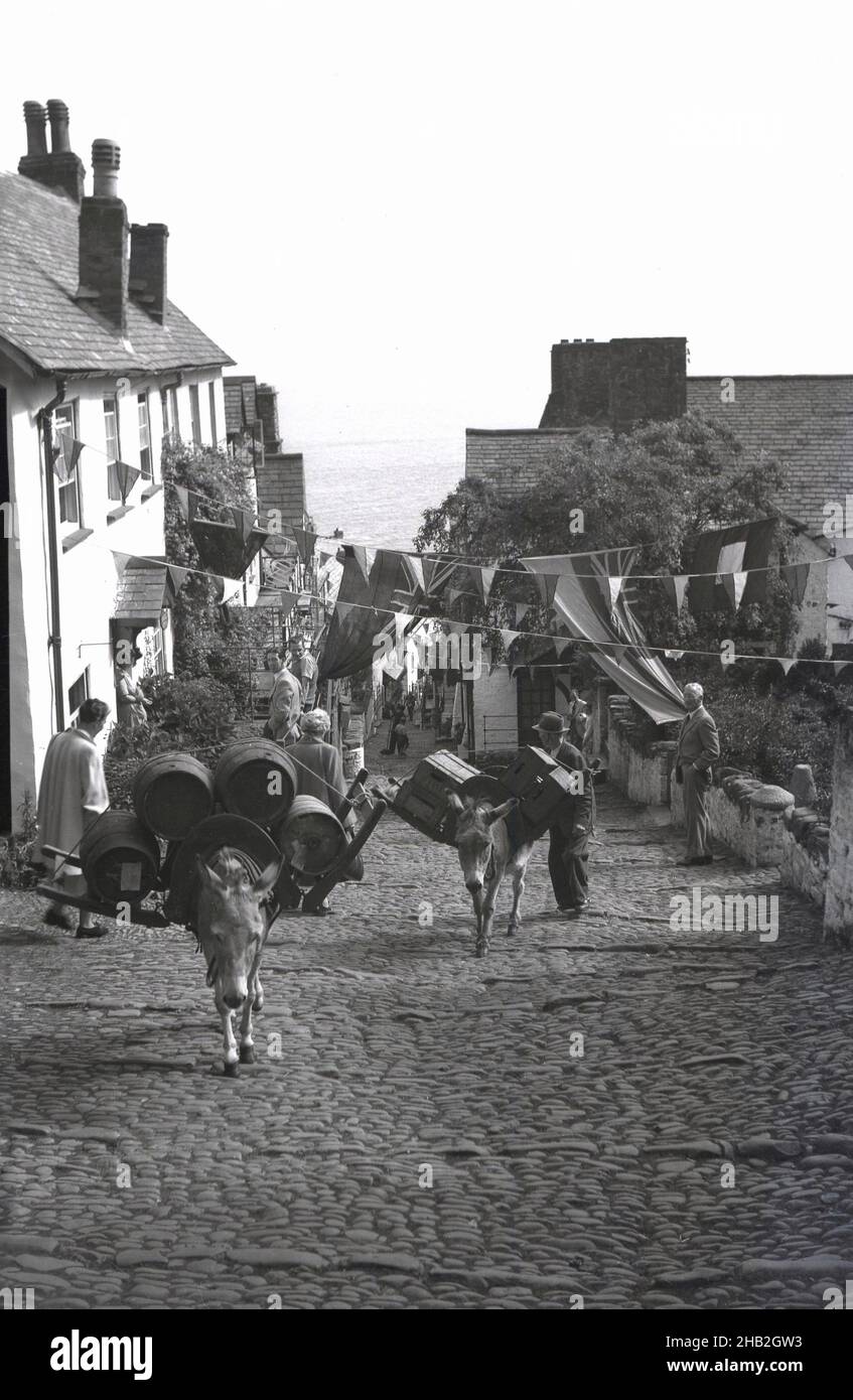 1950s, historisch, zwei arbeitende Esel, die Holzfässer und Kisten mit abgefülltem Bier die steile gepflasterte Straße vom Hafen unten in Clovelly, Devon, England, Großbritannien, hinauftragen. Das Dorf ist berühmt für seine Fußgängerzone, die abfallende gepflasterte Hauptstraße, die traditionellen Hütten und die Architektur und die Esel. Es liegt 400 Meter über dem Hafen von Bideford Bay. Ungewöhnlich ist, dass das Dorf in Privatbesitz ist, da es seit 1738 in der Familie Hamyln lebt. Stockfoto