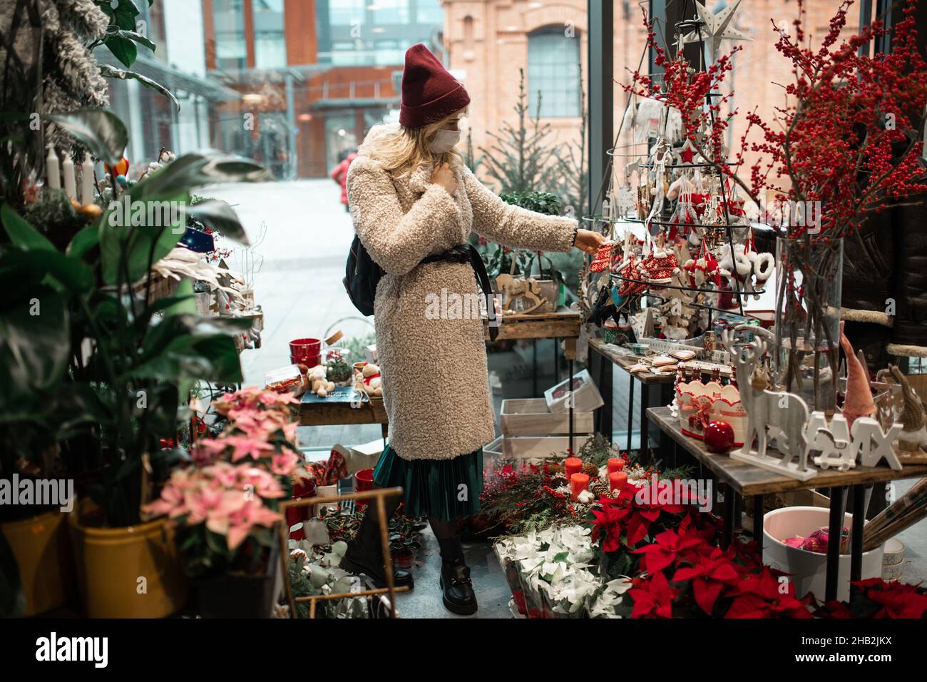Junge Frau in medizinischen Schutzmaske in Kunstpelzmantel und Hut zwischen roten Weihnachtsdekor wählt Geschenke im Blumenladen. Silvester. Weihnachtsdekor Stockfoto