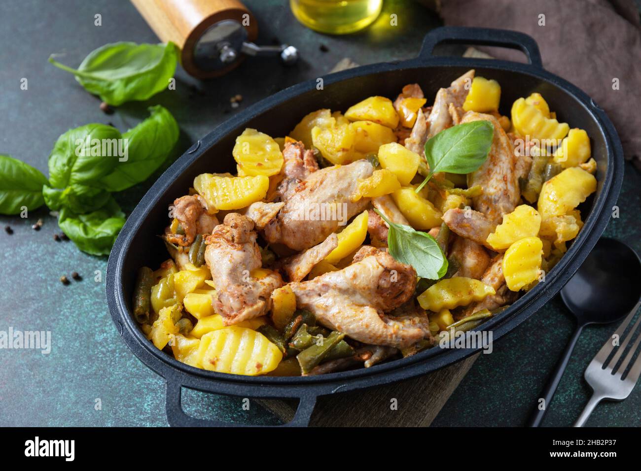 Gebackene Hähnchenflügel mit Kartoffeln, Bohnen, Kräutern, Knoblauch und Bazil auf einer Pfanne auf einer Tischplatte aus dunklem Stein. Hausgemachtes Essen. Griechische und mediterrane Küche. Stockfoto